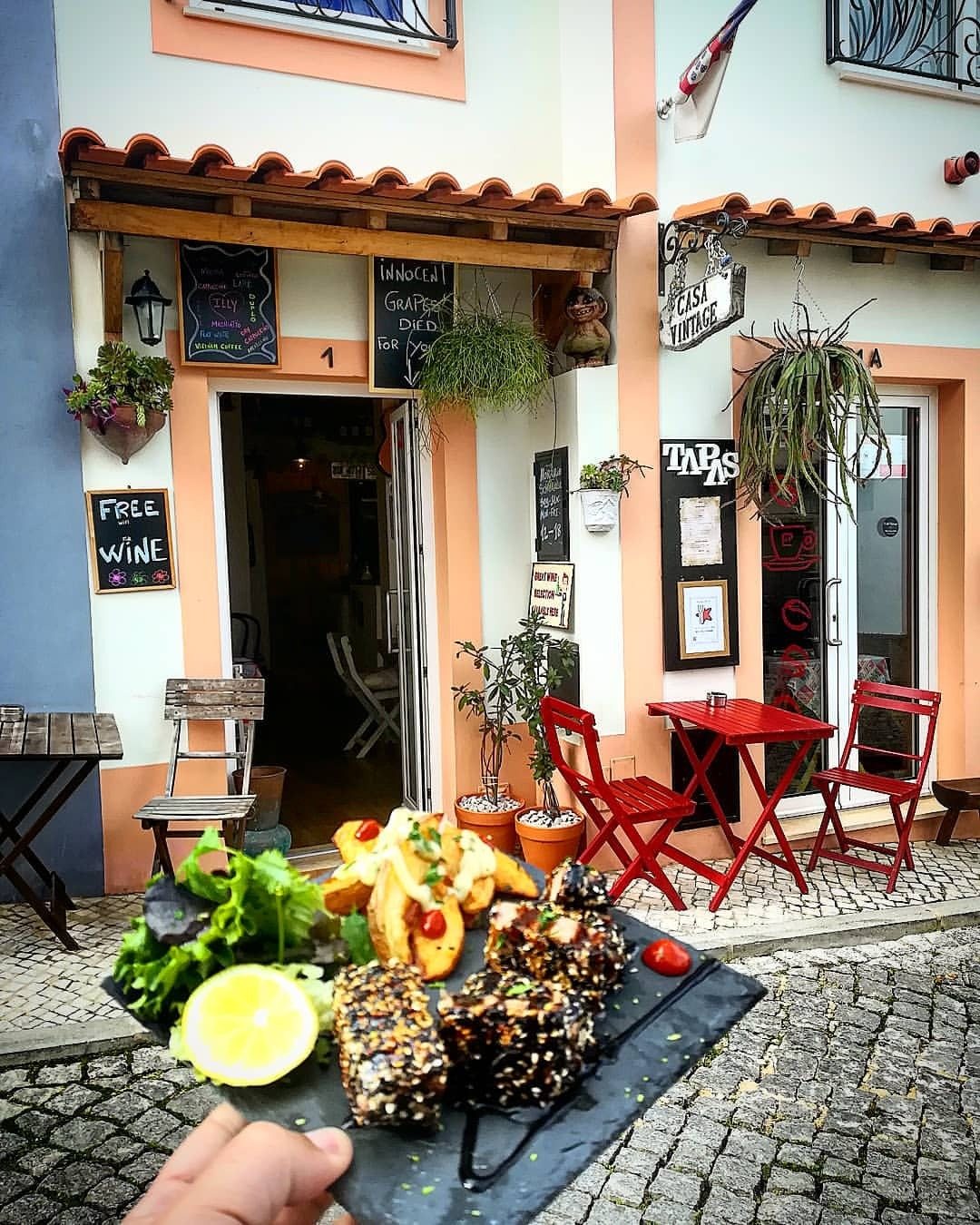 Fachada do Café Vintage numa rua calcetada, com mesas vermelhas à porta e uma travessa de petiscos em primeiro plano
