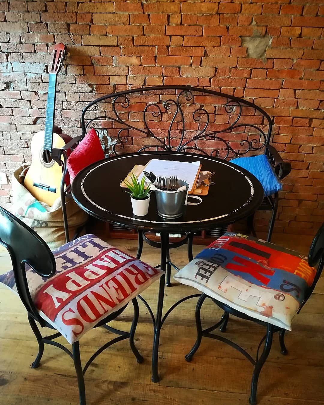 Canto da sala com mesa redonda preta, cadeiras de ferro com almofadas e parede de tijolo